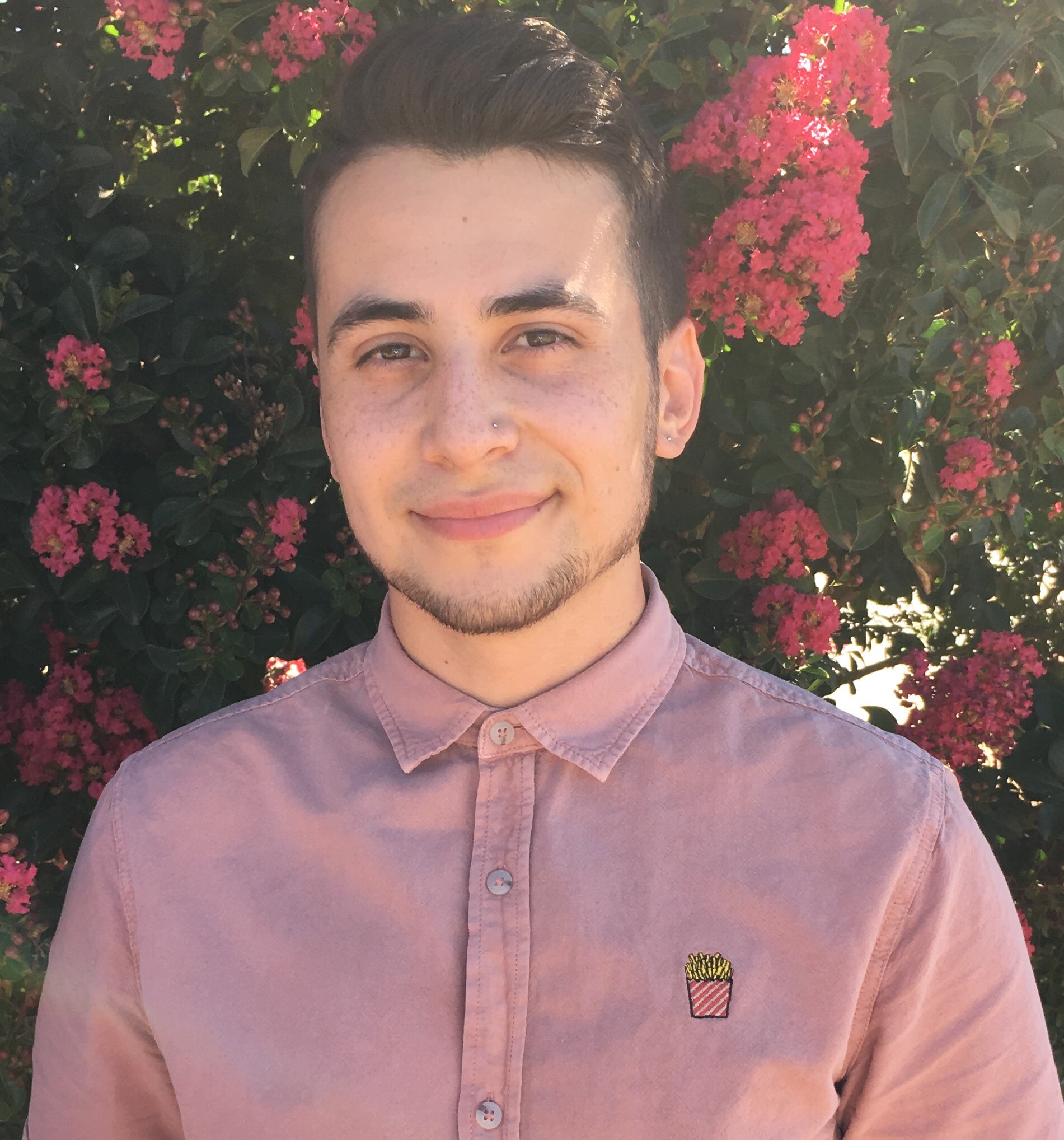 Head and shoulders photo of Angelo with a dusty rose button up top, and a small smile, with a crepe myrtle tree in the background
