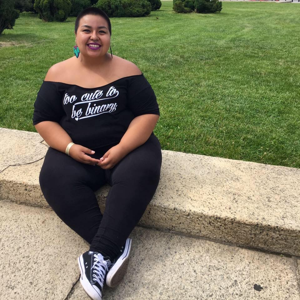 Joanna is seated on a low concrete step with their ankles crossed. They are wearing a shirt with script reading "Too Cute to Be Binary" and they have colorful dangly earrings, bright fuchsia lipstick, snakebite and septum piercings and closely shaved hair.