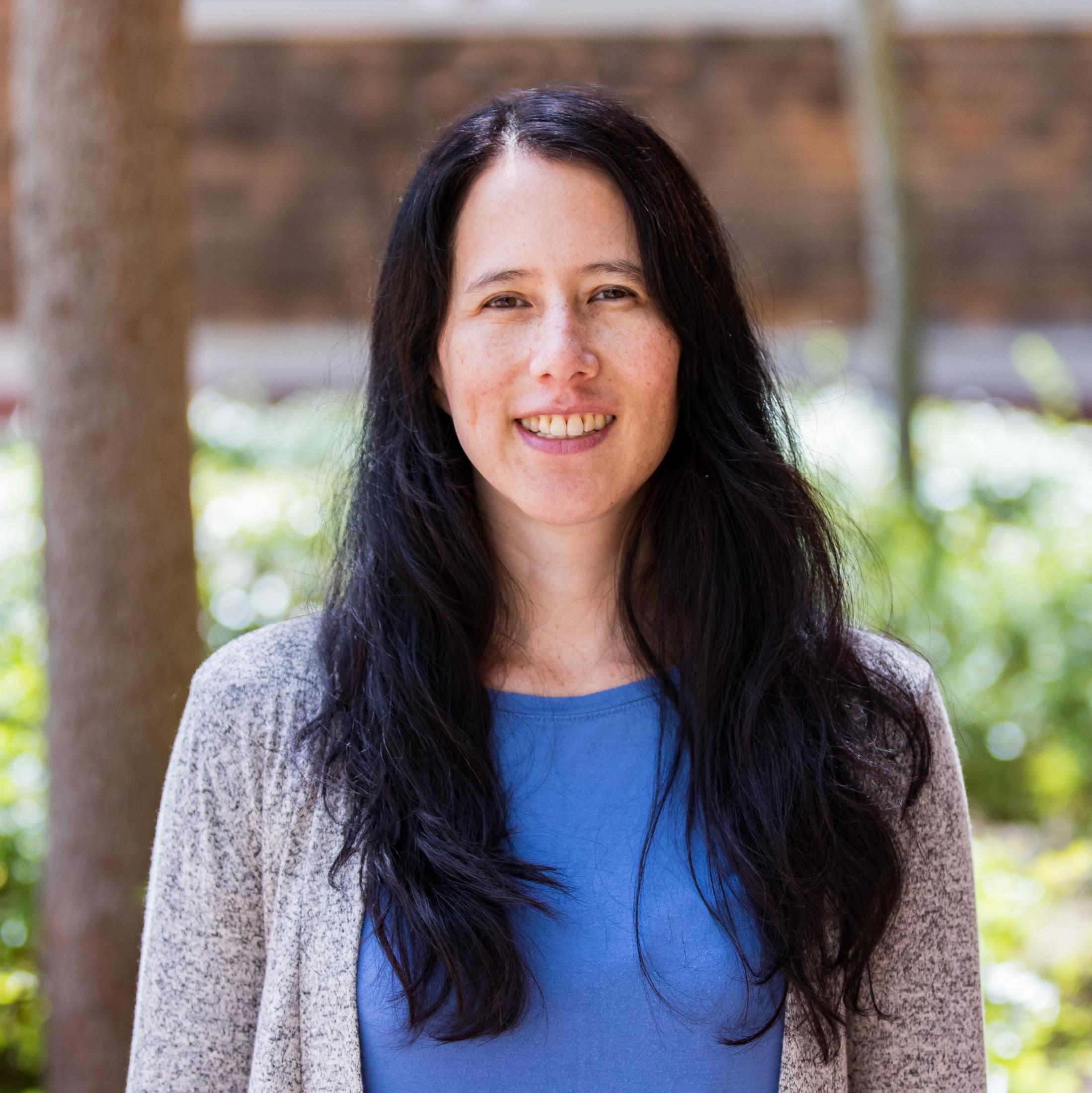 Image of Maia standing, looking at the camera and smiling, with trees in the background.