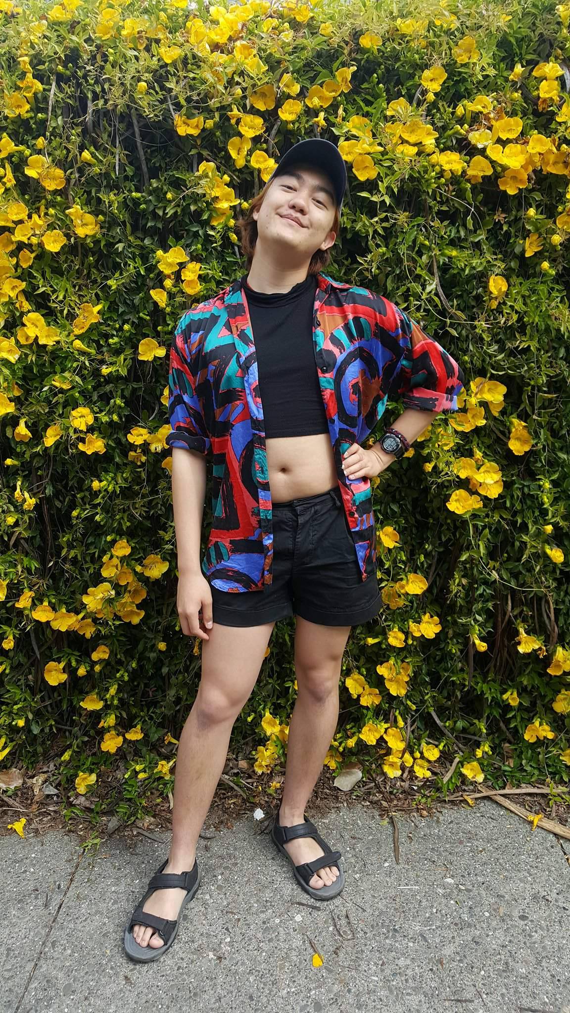 Max standing in front of a yellow flowering shrubbery, with their chin tilted upward and one hand on their hip. Max is wearing shorts, crop top, and graphic print blouse.