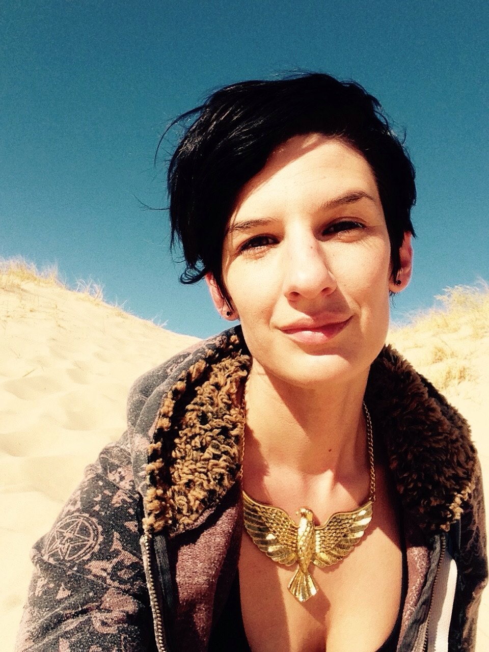 elizabeth coté sitting in Mojave sand dunes, smiling, wearing a golden hawk necklace.