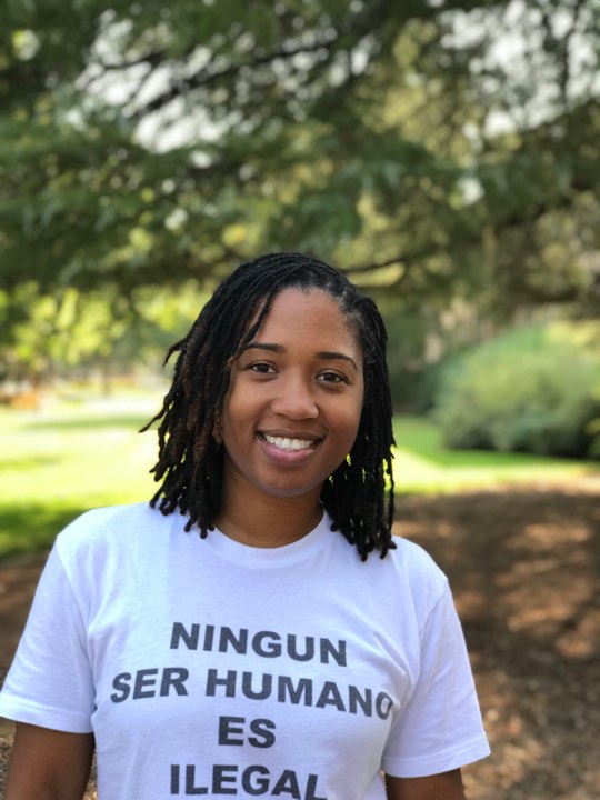 Head and shoulders photo of Monae. Monae is smiling and tree branches are in the background. Monae's shirt reads: Ningun ser humano es ilegal.