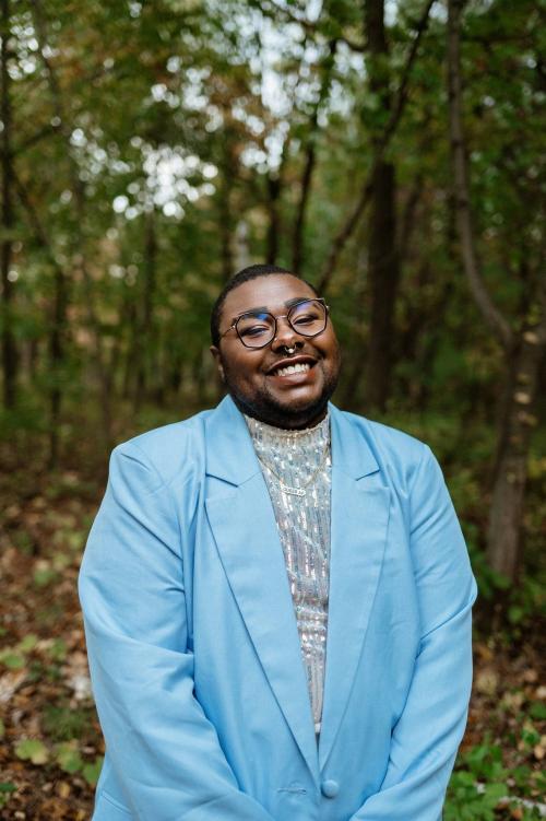 TK, a black trans and queer person, is wearing a light blue suit and glasses and smiling at the camera in front of a woody landscape