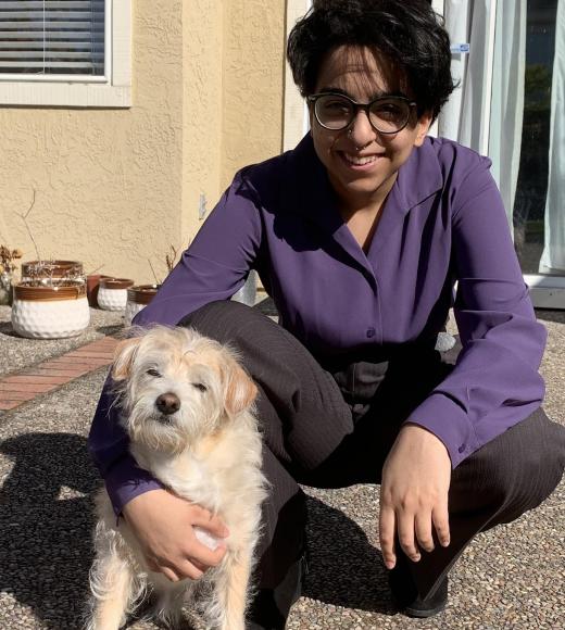 Meghan is bespectacled, has brown skin and black short hair. they are wearing black slacks and a purple dress shirt. she is lovingly holding a small brown dog.
