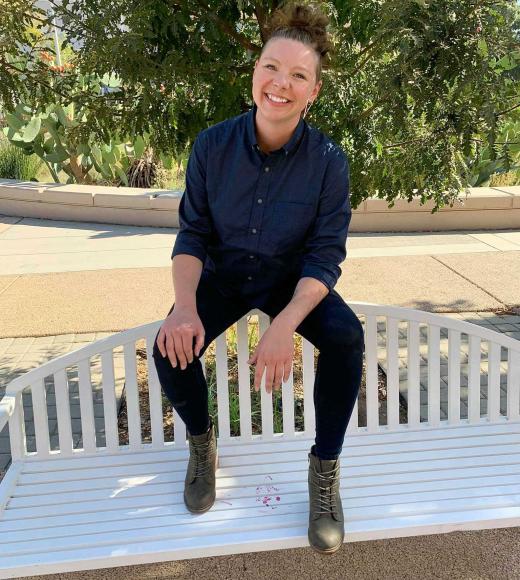 Crystal is a white tall person with blonde hair in a bun and shaved sides. they are smiling at the camera while posed on a white bench in a blue button down and black skinny jeans with boots.