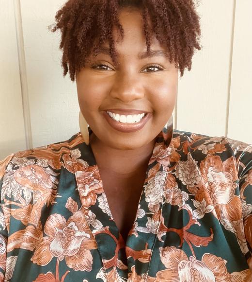 Talia, a Black Queer femme, is smiling warmly at the camera. She is wearing a multi-color floral shirt and has a bright smile and auburn curly bangs