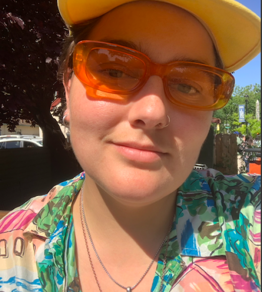 Jasper, a white person, is smiling at the camera. They are in the sun in orange sunglasses and a bright floral shirt with a yellow garfield cap.