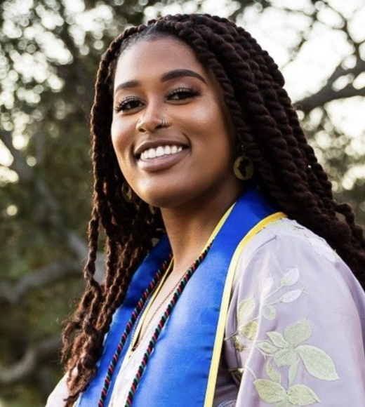 graduation photo of Zi smiling with a blue and gold stole and long hair, with green trees in the background