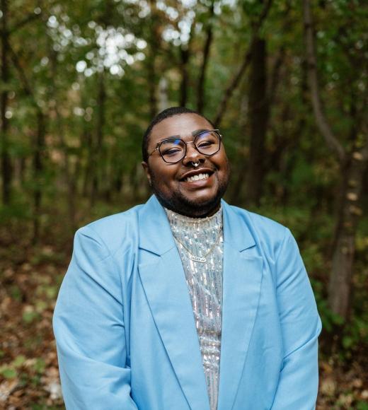 TK, a black trans and queer person, is wearing a light blue suit and glasses and smiling at the camera in front of a woody landscape