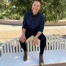 Crystal is a white tall person with blonde hair in a bun and shaved sides. they are smiling at the camera while posed on a white bench in a blue button down and black skinny jeans with boots.