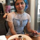 Echo has brown skin, blue hair, and very cute makeup. they are wearing a blue dress over a white top and are smiling over a plate of waffles. 