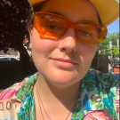 Jasper, a white person, is smiling at the camera. They are in the sun in orange sunglasses and a bright floral shirt with a yellow garfield cap.