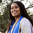 graduation photo of Zi smiling with a blue and gold stole and long hair, with green trees in the background