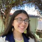 La'a is wearing glasses, has tan skin, long black hair, and a big smile. he is looking at the camera warmly and wearing fun earrings and a blue and cream windbreaker over a pale yellow tee shirt. 